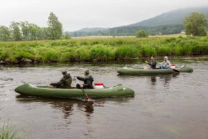 vodáci na Vltavě, v popředí jsou vidět dvě zelené kánoe, ve kterých sedí lidé s pádly, na břehu řeky je hustý travní porost a louka